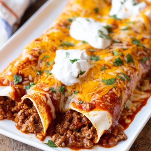 Tender flour tortillas stuffed with seasoned ground beef and cheddar cheese, smothered in vibrant red enchilada sauce for a classic Mexican dinner.
